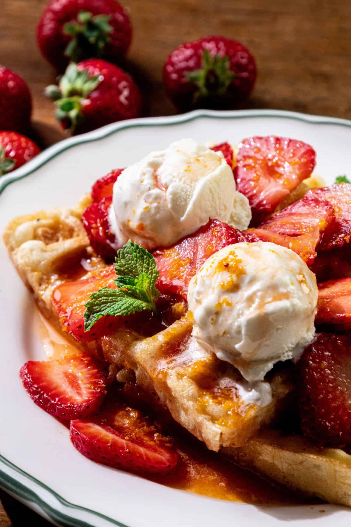 Waffles with macerated strawberries, vanilla ice cream, sauce drizzle, mint sprig, and whole strawberries in the background.