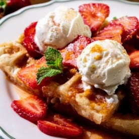 Waffles with strawberries, vanilla ice cream scoops, a mint sprig, and orange zest on a white plate.