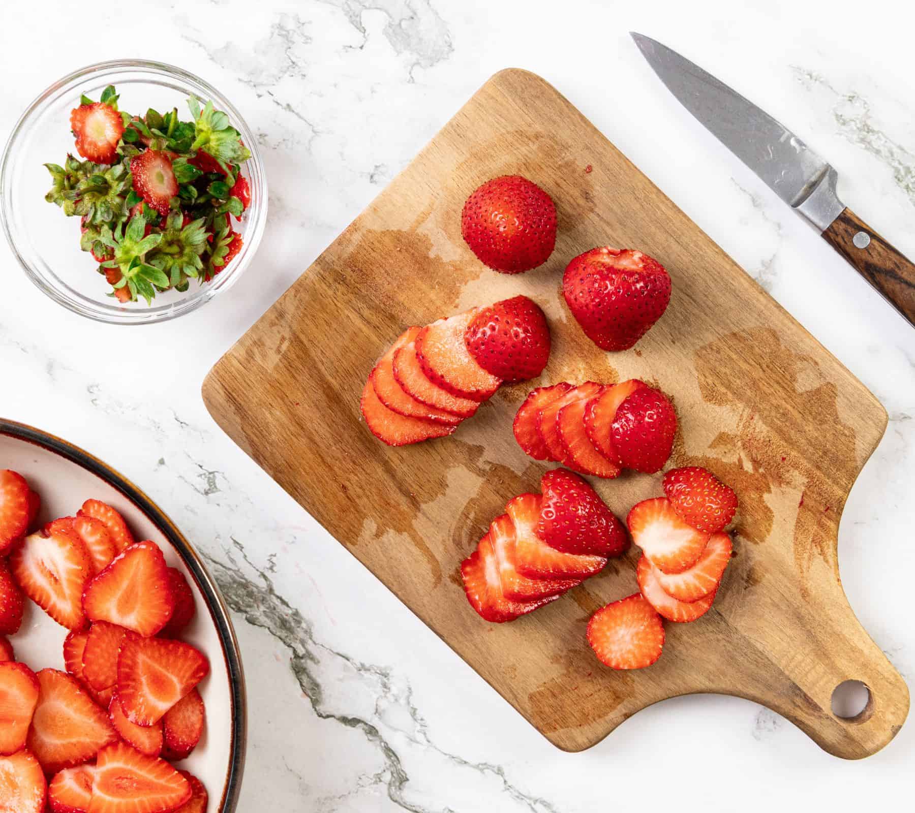 Sliced strawberries on a wooden cutting board, a knife, a bowl of macerated strawberries, and a jar of strawberry tops.