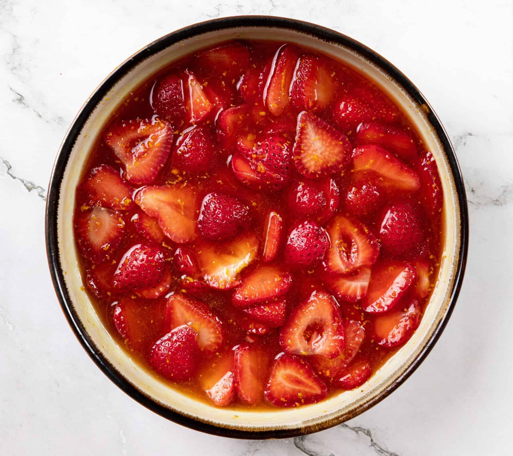 A bowl of strawberries in glossy red syrup on a white marble surface.