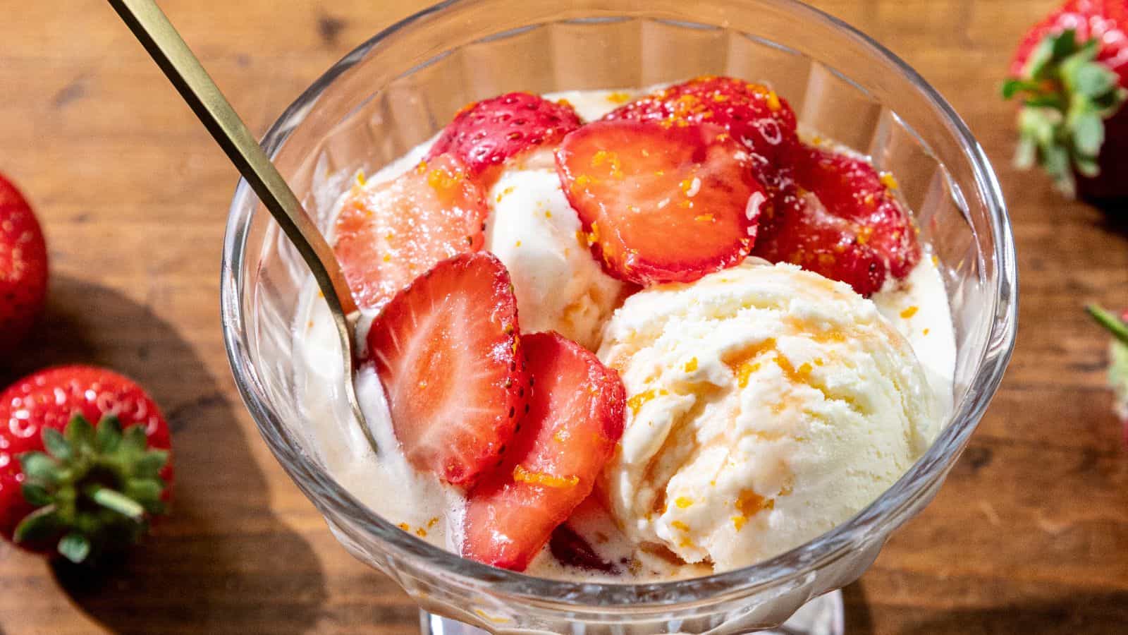 Glass bowl of vanilla ice cream with boozy strawberries on top, a spoon to the left, all placed on a wooden table.