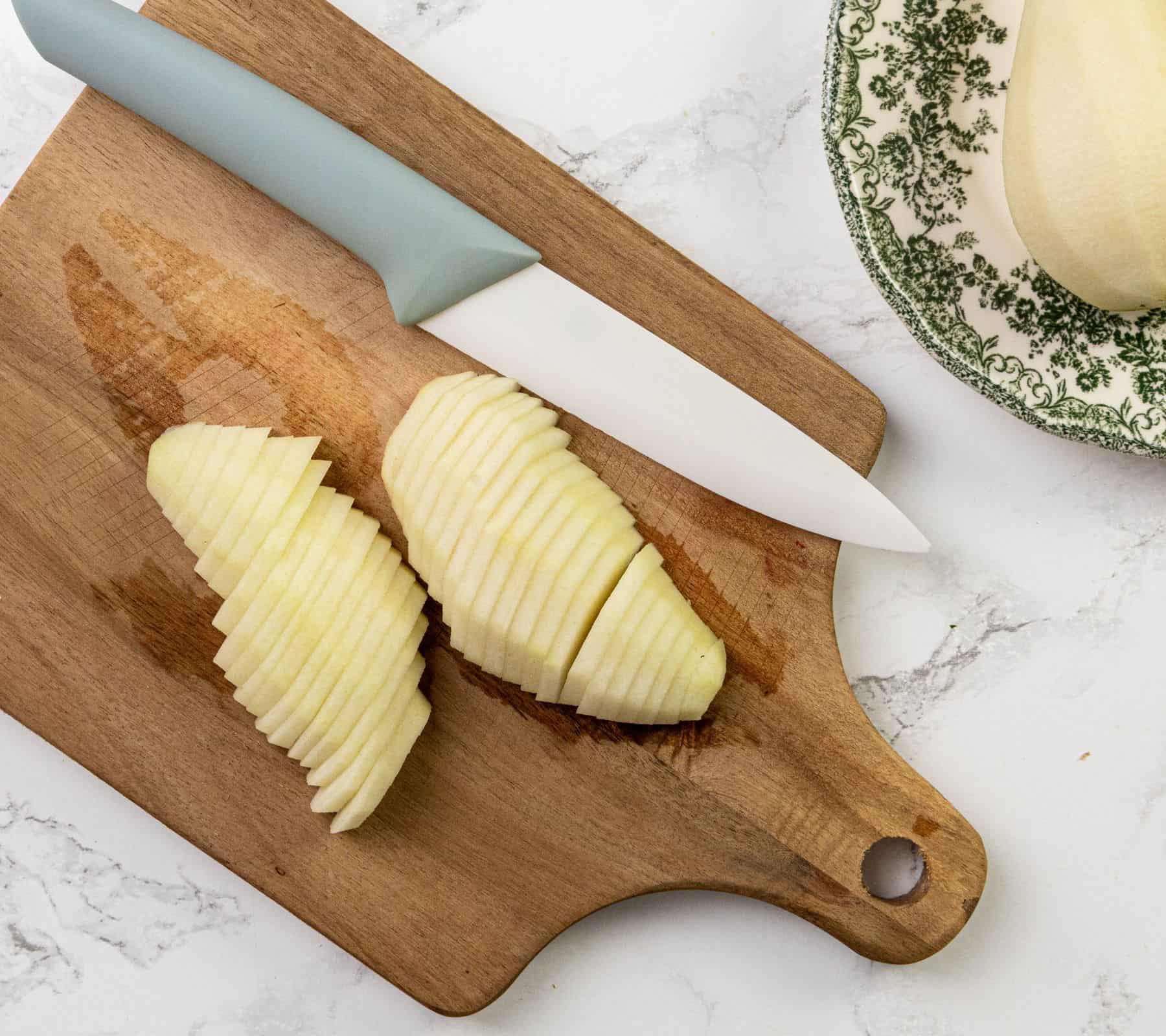 A ceramic knife and two peeled, thinly sliced apples on a wooden cutting board with part of a patterned plate nearby.