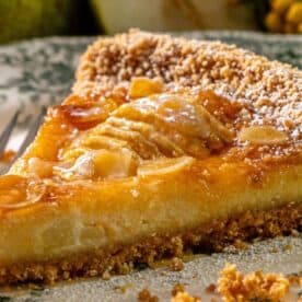 A slice of pear tart with crumbly topping and glossy glaze rests on a decorative plate next to a fork.