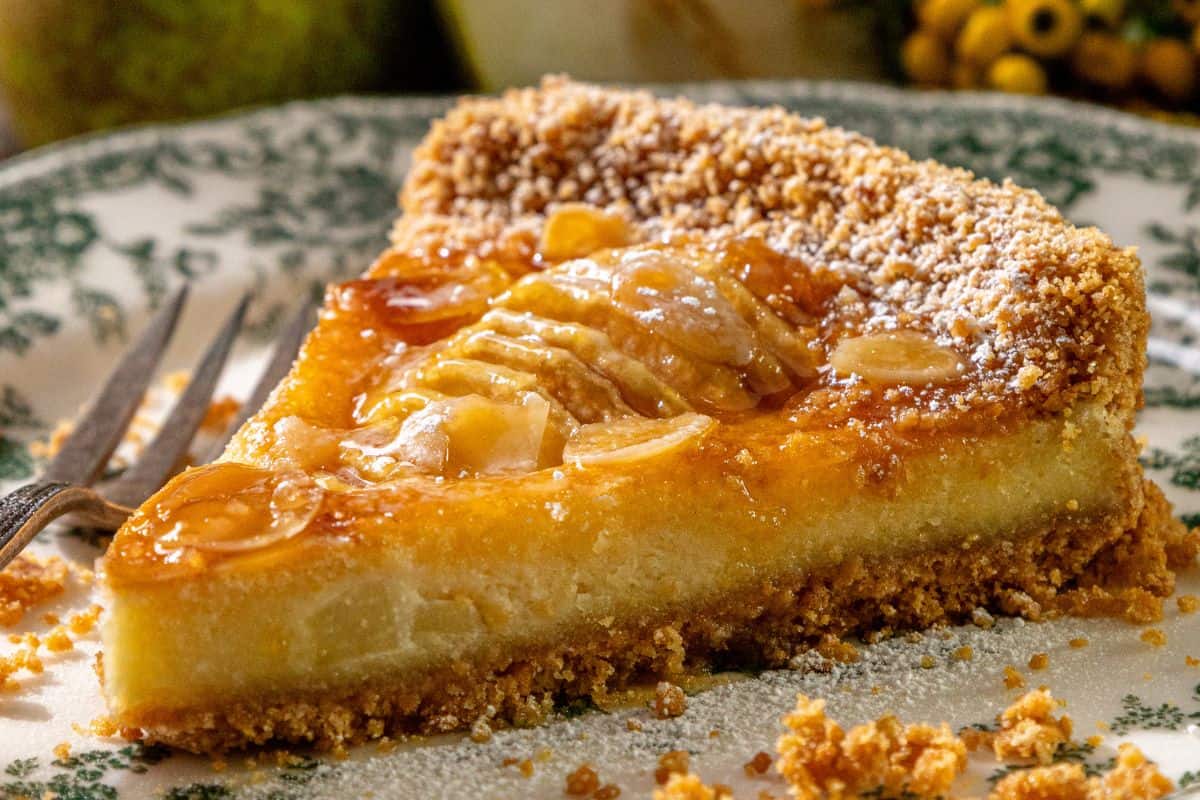 A slice of pear tart with crumbly topping and glossy glaze rests on a decorative plate next to a fork.