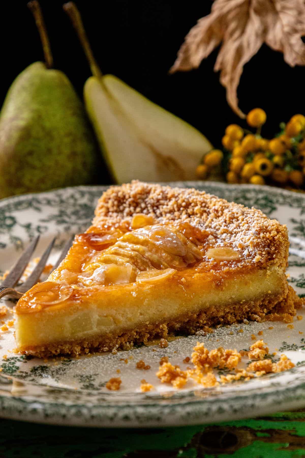 A slice of pear tart with golden shortbread crust on a decorative plate, with pears and yellow berries in the background.