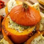 A baked pumpkin with its top off, filled with creamy herbs, sits on a platter with sliced pumpkin pieces around it.