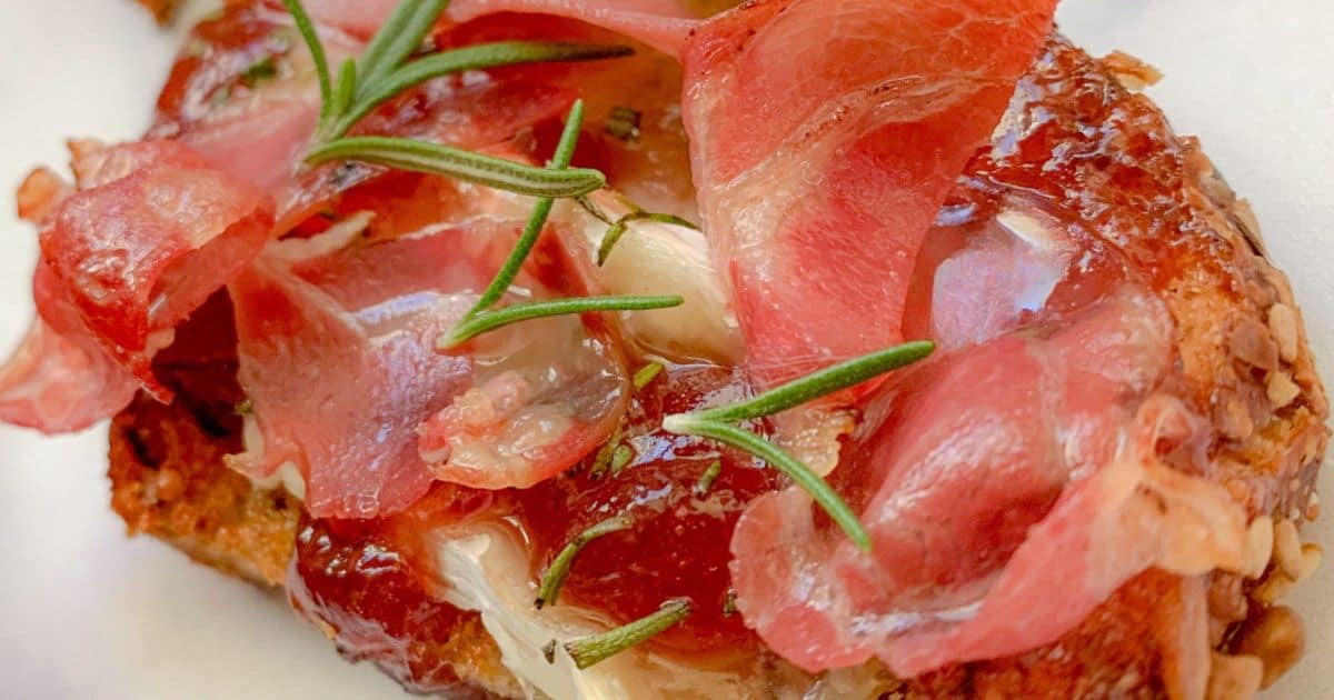 Close-up of a slice of toasted bread topped with thin slices of cured meat and sprigs of fresh rosemary, glistening with a drizzle of oil. The dish is presented on a white background, showcasing its vibrant textures and colors.