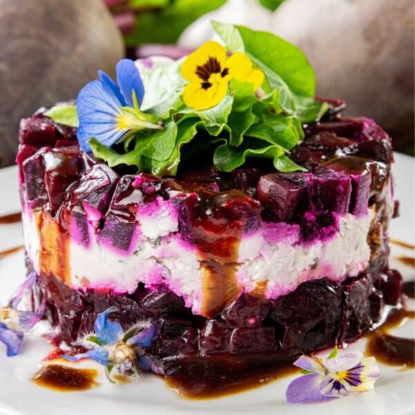 A layered salad featuring diced beets and cheese, topped with edible flowers and fresh greens, finished with a dark balsamic drizzle, presented on a white plate.
