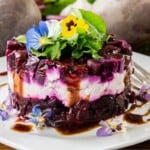 A layered salad of diced beets, cheese, dark sauce, and edible flowers sits on a white plate with a fork. Whole beets are visible in the background.
