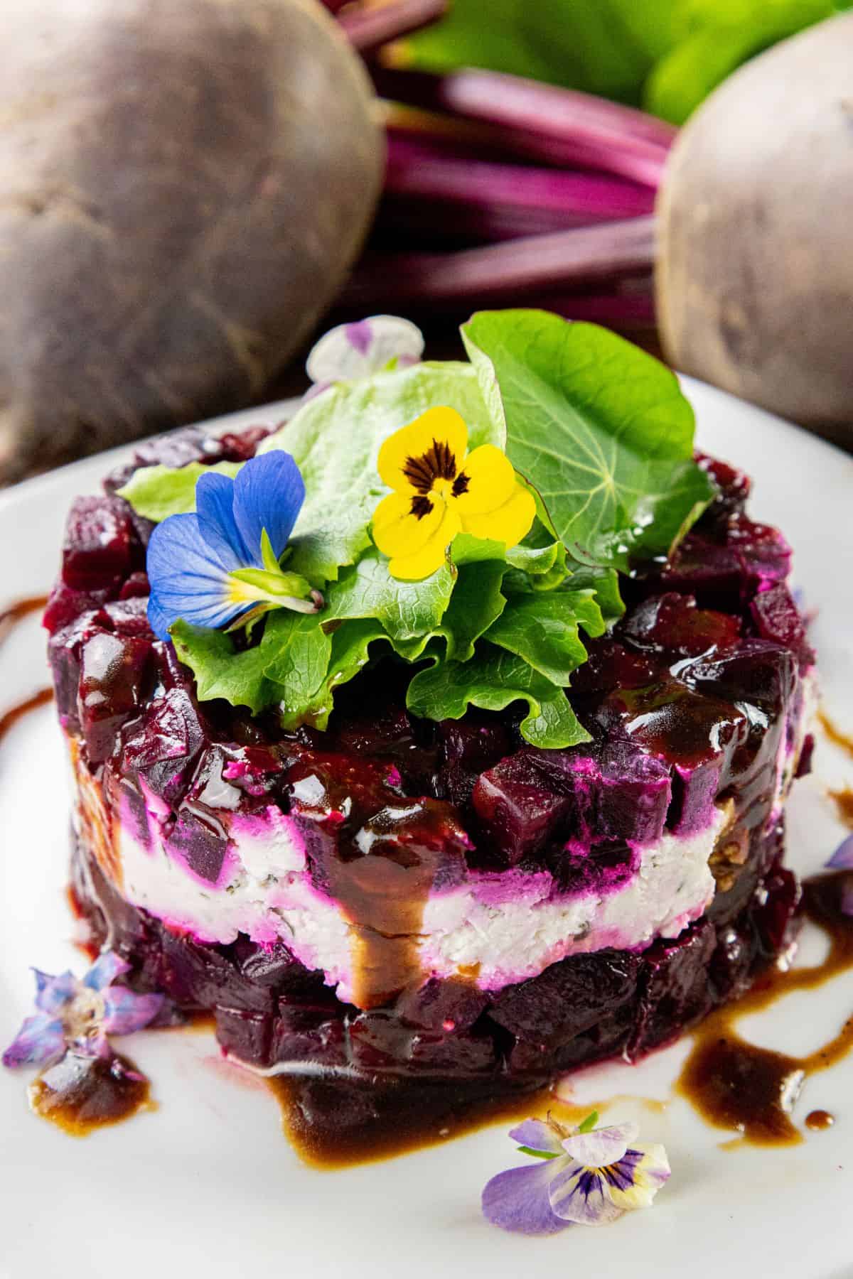 The image shows a layered stack of sliced beets and cheese, garnished with edible flowers and fresh greens, arranged on a white plate. In the background, whole beets are visible.