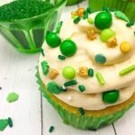 Cupcake in green wrapper with white frosting, green and gold sprinkles, green candies on white surface; bowl of green sprinkles behind.