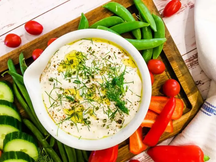 A bowl of creamy dip garnished with herbs and olive oil is placed on a wooden tray. The tray also contains green beans, snap peas, cucumber slices, red bell peppers, and cherry tomatoes, creating a vibrant and colorful presentation.