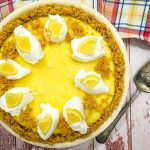 A lemon pie with whipped cream dollops and lemon wedges atop a crumbled cookie crust sits on a wooden surface alongside a colorful checkered cloth and a metal pie server.