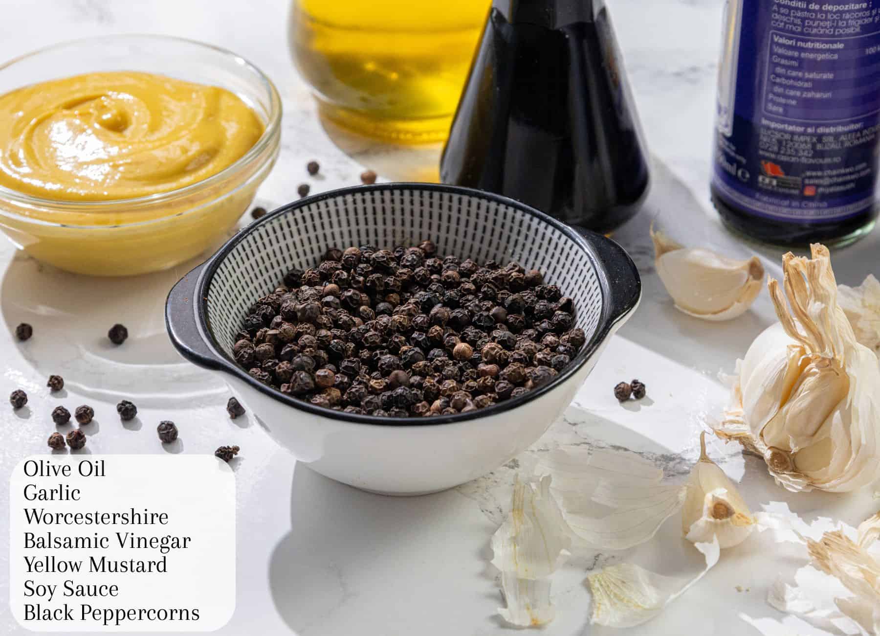 A bowl of black peppercorns, garlic cloves, yellow mustard, balsamic vinegar, oil and sauce bottles on a white surface. A printed ingredient list lies in front.
