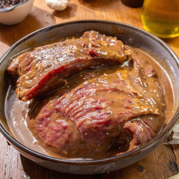Two pieces of raw beef in a round bowl with marinade on a wooden surface.