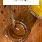 A jar of brown steak marinade with a spoon sits on a wooden surface beside a bowl of yellow liquid and scattered peppercorns.
