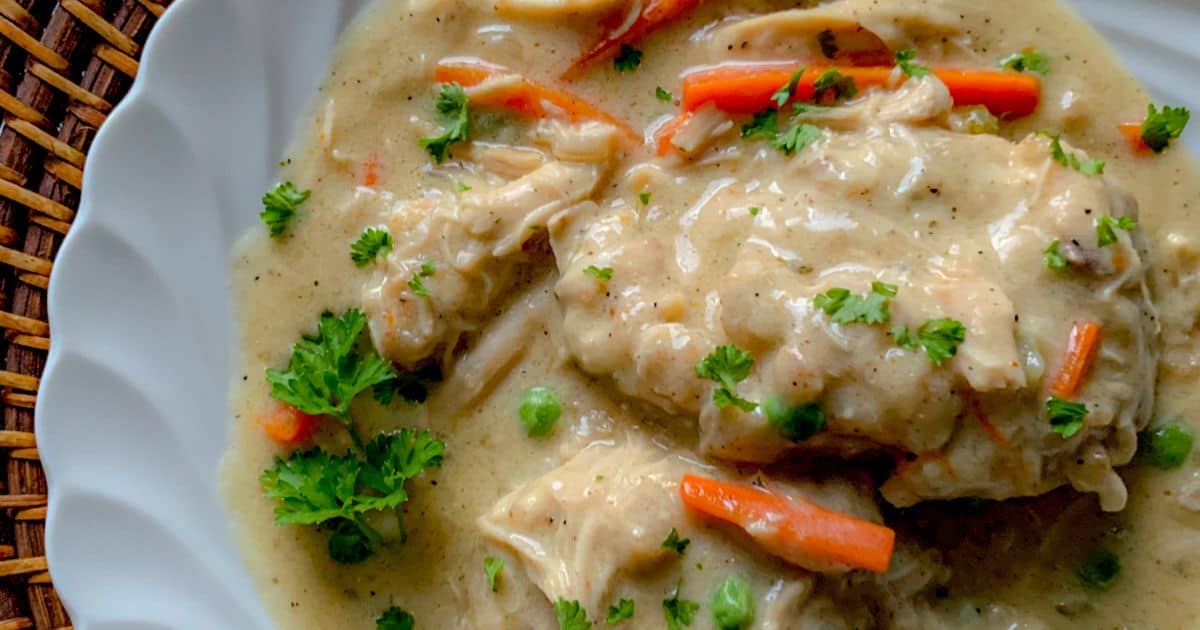 A close-up of creamy chicken and dumplings garnished with parsley. The dish includes pieces of chicken, dumplings, sliced carrots, and peas in a thick, savory sauce, served on a white plate with a woven placemat underneath.
