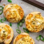 On a baking tray, there are six cheese puff pastry appetizers with visible herbs, garnished with chopped green onions.