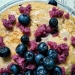 A large vanilla cake on a plate, with blueberries and blueberry sugar.