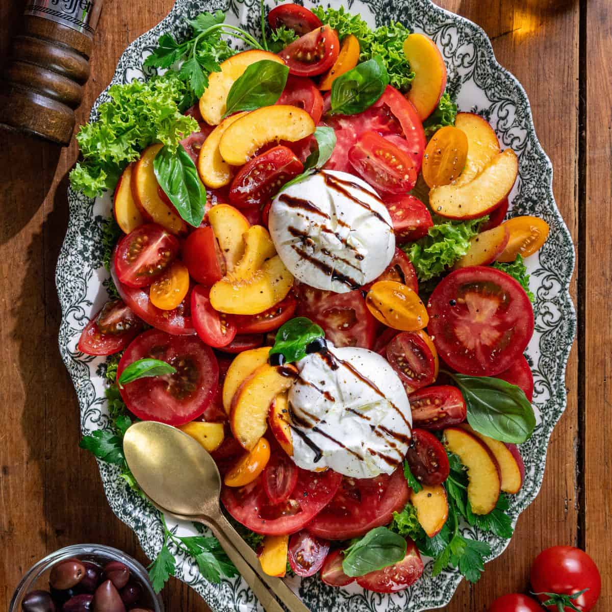 A platter with sliced tomatoes, peaches, lettuce, basil leaves, mozzarella balls topped with balsamic glaze, and a gold serving spoon on the side.