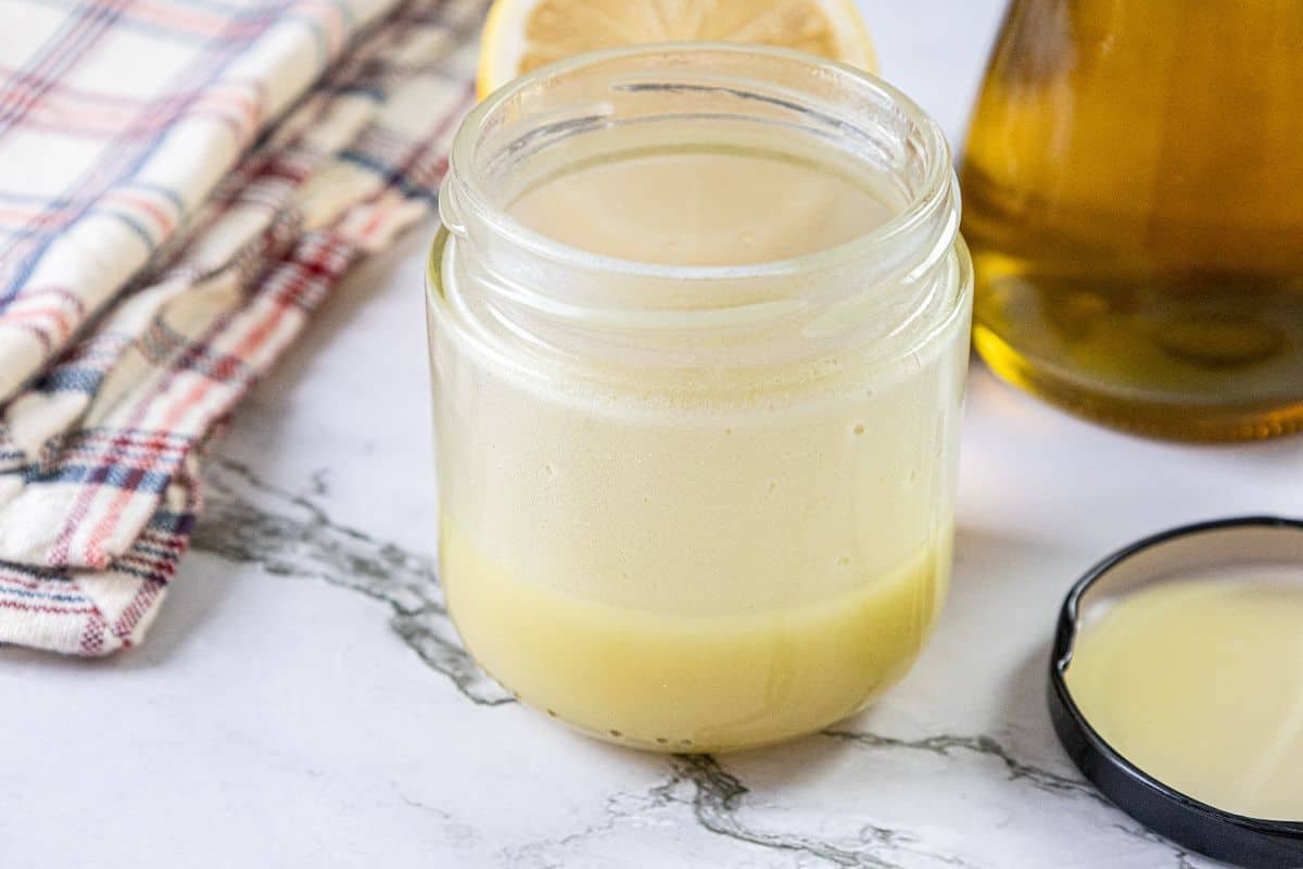 An open glass jar filled with light yellow liquid sits on a marble surface beside a jar lid, a plaid cloth, a lemon slice, and a bottle.
