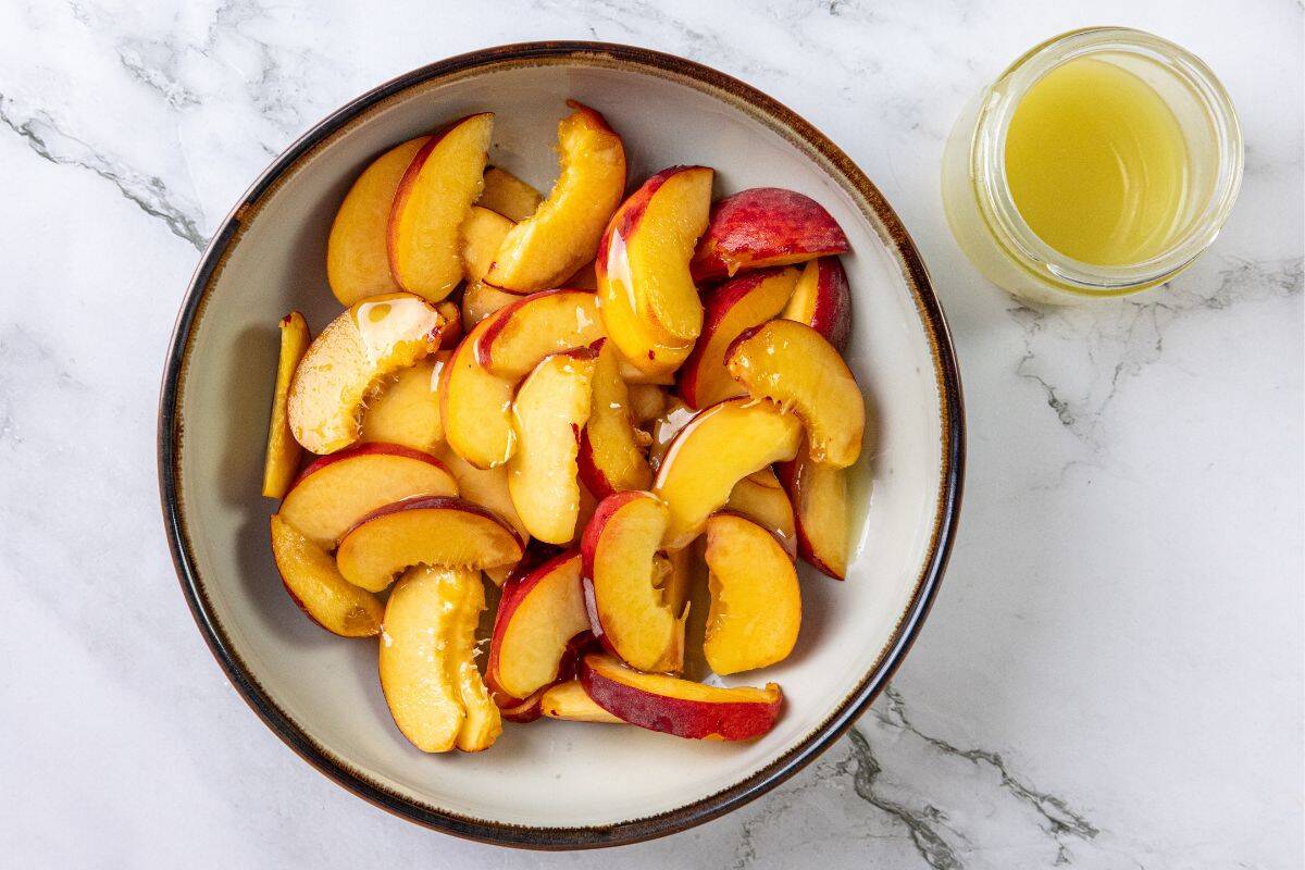 A bowl of sliced peaches and a glass jar filled with light yellow liquid rest on a white marble surface.