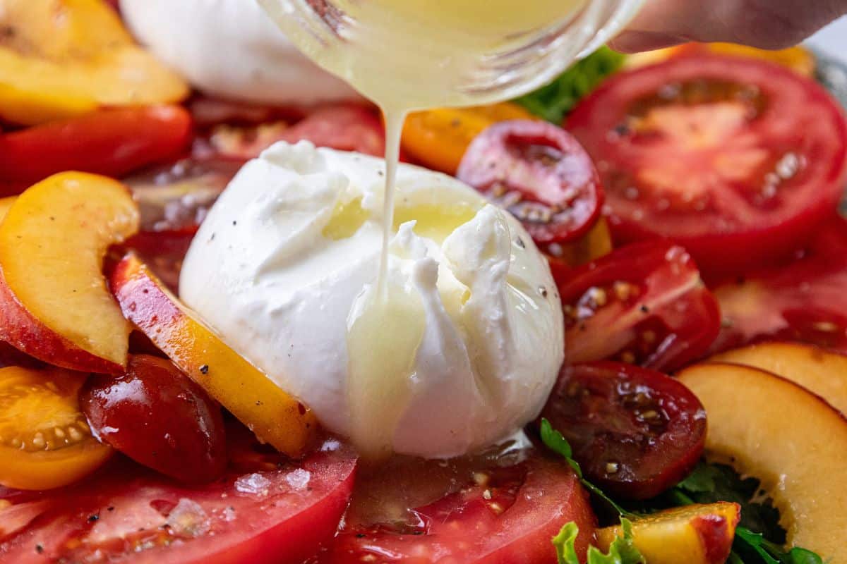 Burrata cheese, sliced tomatoes, and peaches arranged together, with a light dressing being poured over the salad.