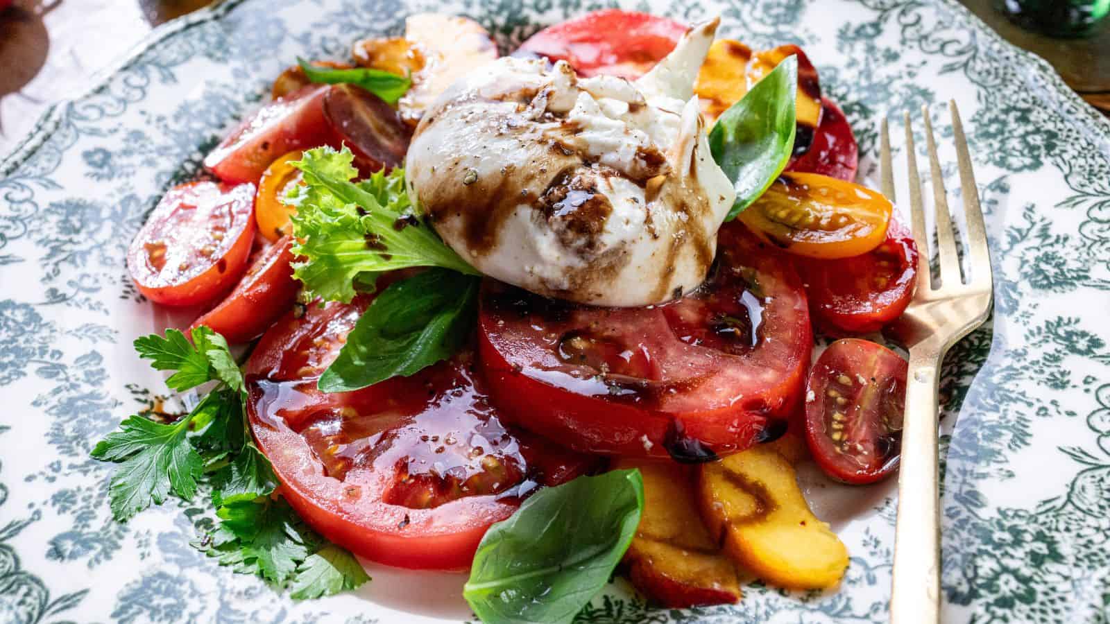 Sliced tomatoes, peaches, and creamy burrata cheese topped with balsamic glaze and fresh basil, arranged on mixed greens on a decorative plate with a fork.