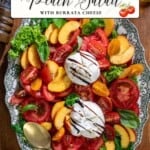 A platter arranged with sliced peaches, tomatoes, fresh basil leaves, lettuce, and burrata cheese, drizzled with balsamic glaze. Surrounding the platter are loose salad ingredients and a serving spoon.