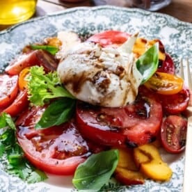 The image shows sliced tomatoes, sliced peaches, leafy greens, burrata cheese, fresh basil leaves, and a drizzle of balsamic glaze on a plate with a fork on the side.