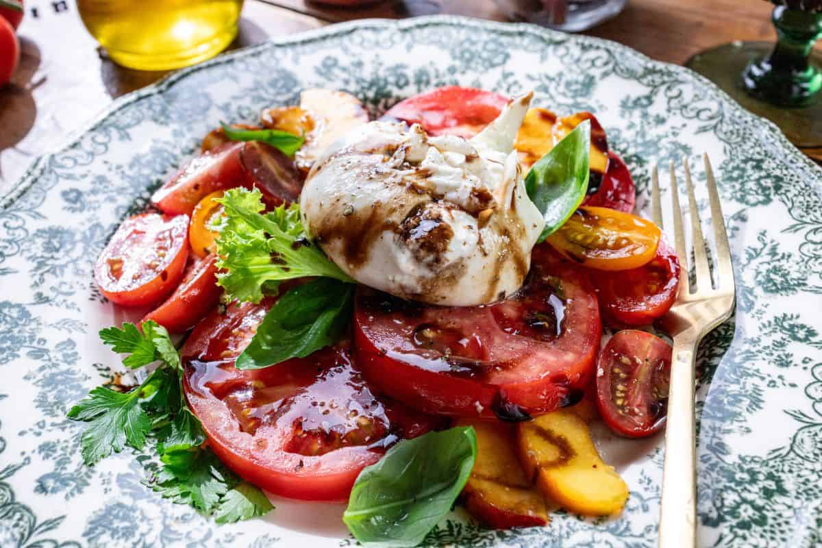 The image shows sliced tomatoes, sliced peaches, leafy greens, burrata cheese, fresh basil leaves, and a drizzle of balsamic glaze on a plate with a fork on the side.