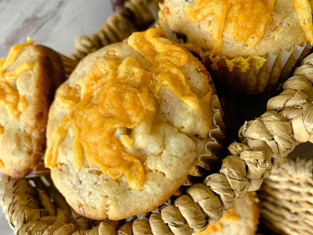 Close-up of a basket containing freshly baked muffins topped with melted cheese. The muffins are in paper liners and have a golden-brown crust. The wicker basket adds a rustic touch to the presentation.