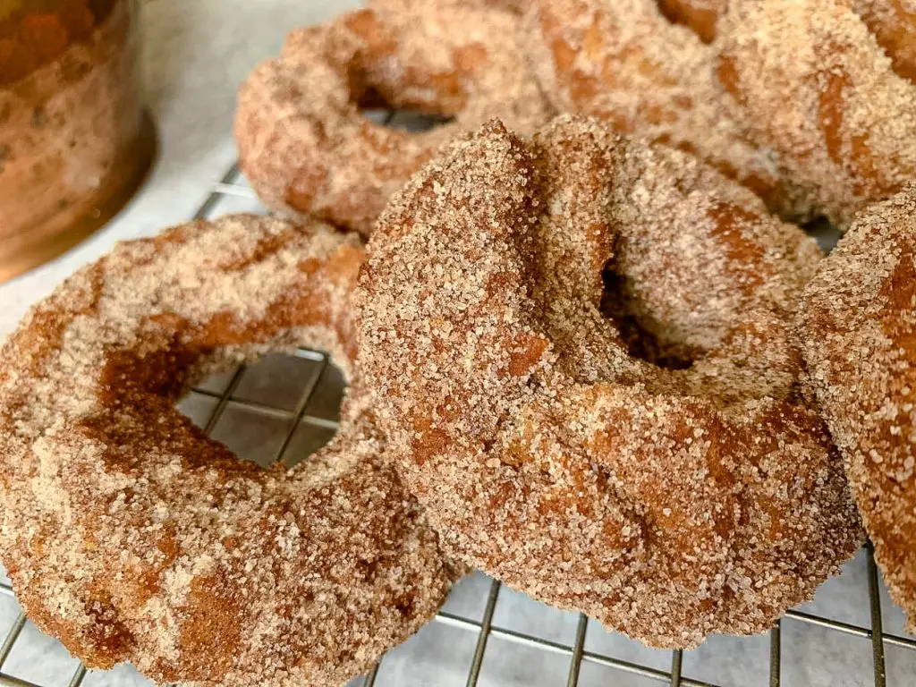 Close-up of several freshly baked donuts covered in a crumbly cinnamon sugar mixture. They are resting on a cooling rack, with a slightly blurred background that includes a hint of a copper pot. The donuts appear golden brown and have a soft texture.