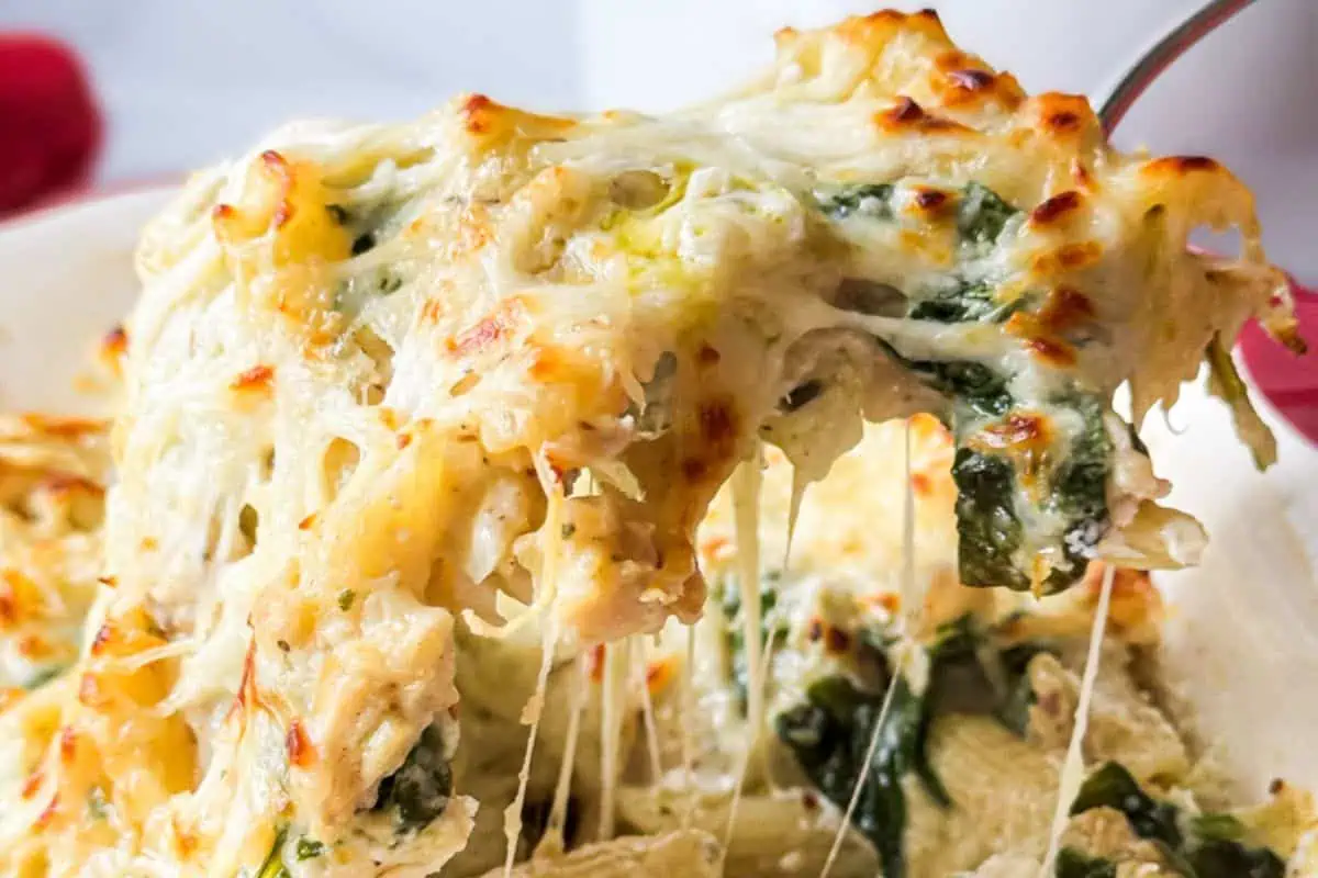 Close-up of a spoon lifting a portion of cheesy, baked spinach artichoke dip, showcasing its melted cheese and creamy texture. The dip includes visible pieces of artichoke and spinach, with a golden-brown layer of cheese on top.
