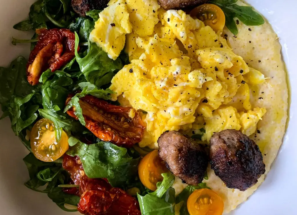 A close-up of a colorful breakfast bowl featuring scrambled eggs, leafy greens, sun-dried tomatoes, cherry tomatoes, and browned sausage pieces. The dish is seasoned with black pepper, adding a touch of flavor to the vibrant and healthy meal.