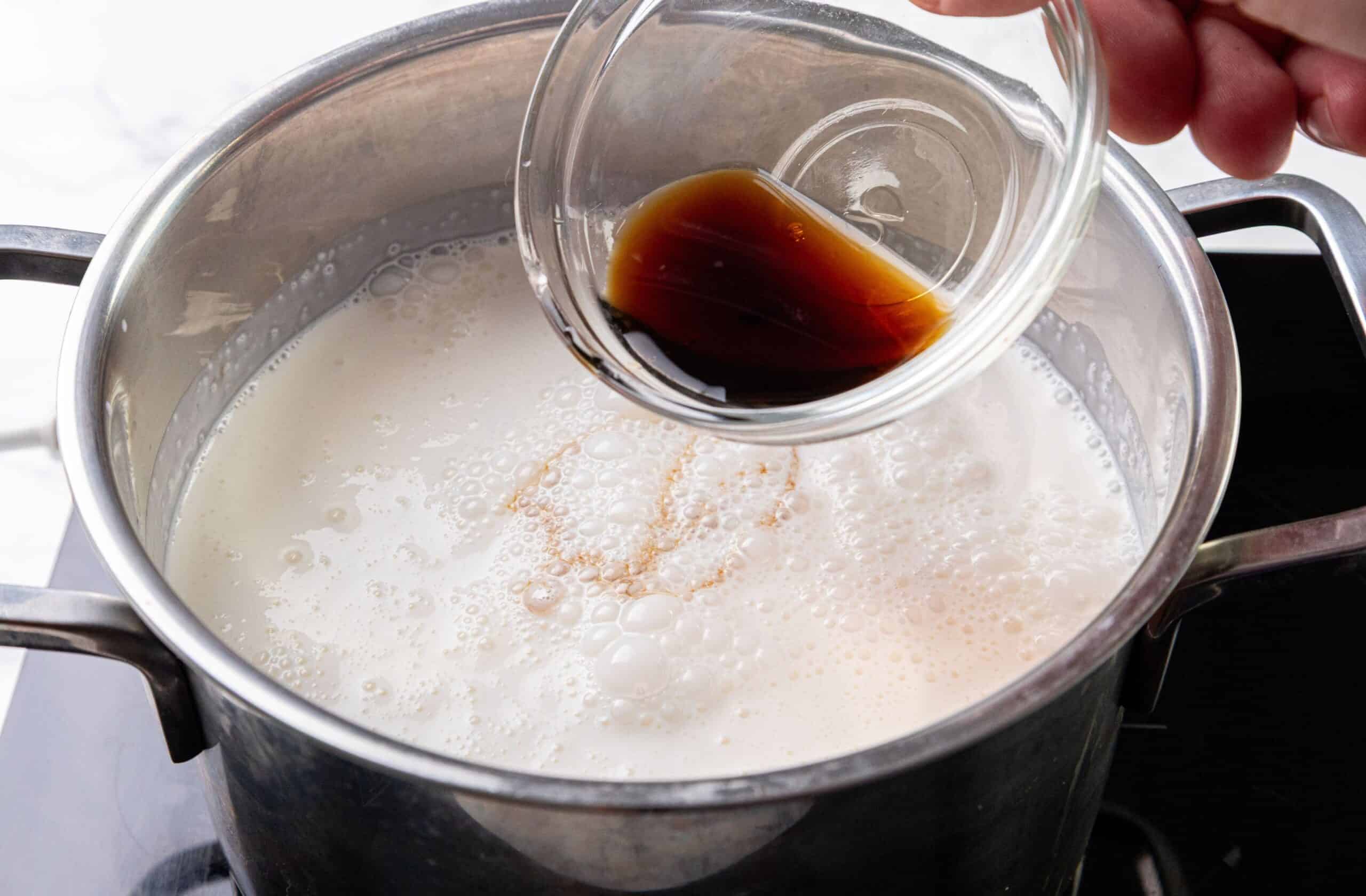 A person pours vanilla extract from a small bowl into a pot of milk on a stovetop.