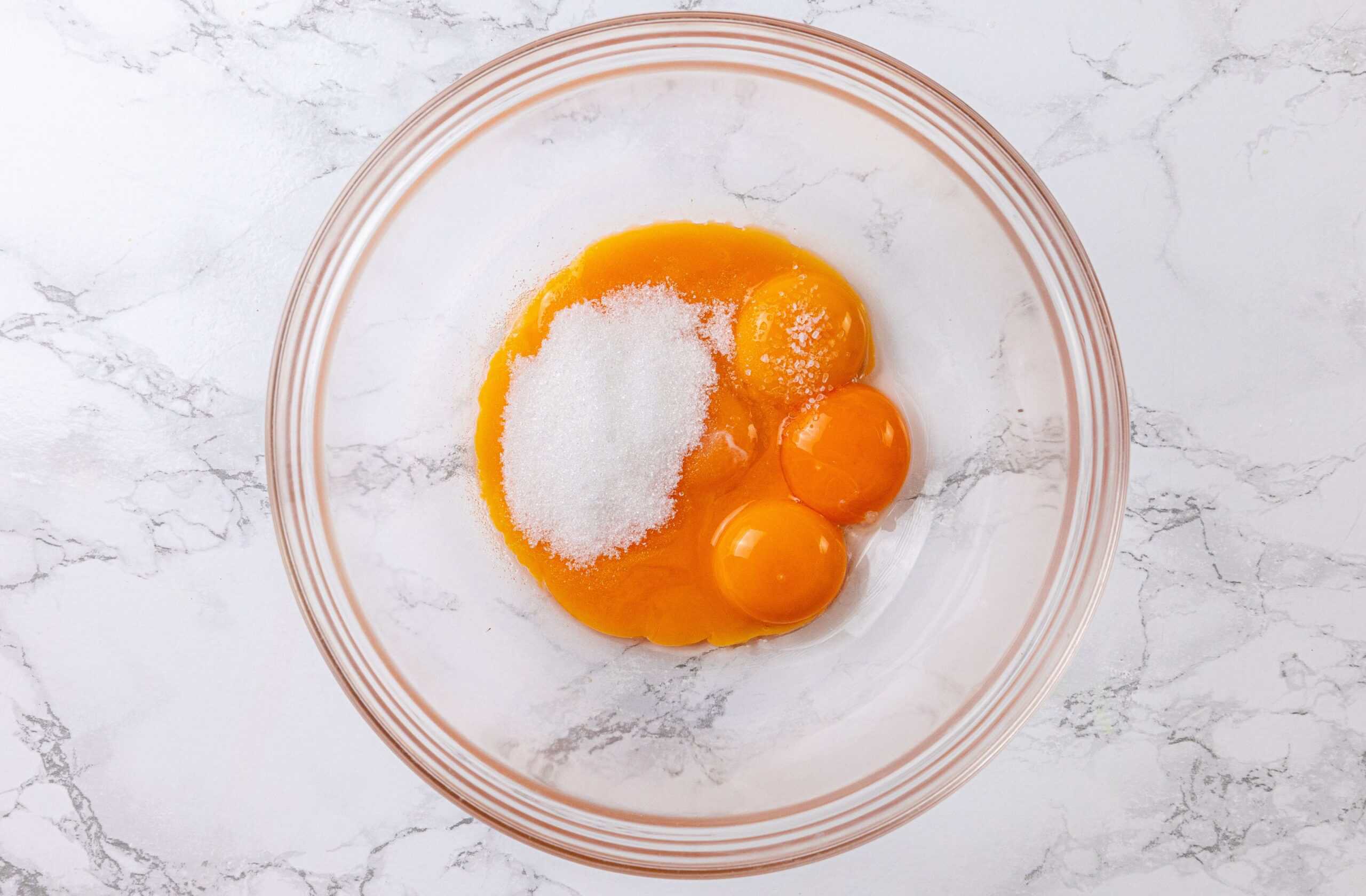 A glass bowl with three egg yolks and a mound of granulated sugar on a white marble surface.