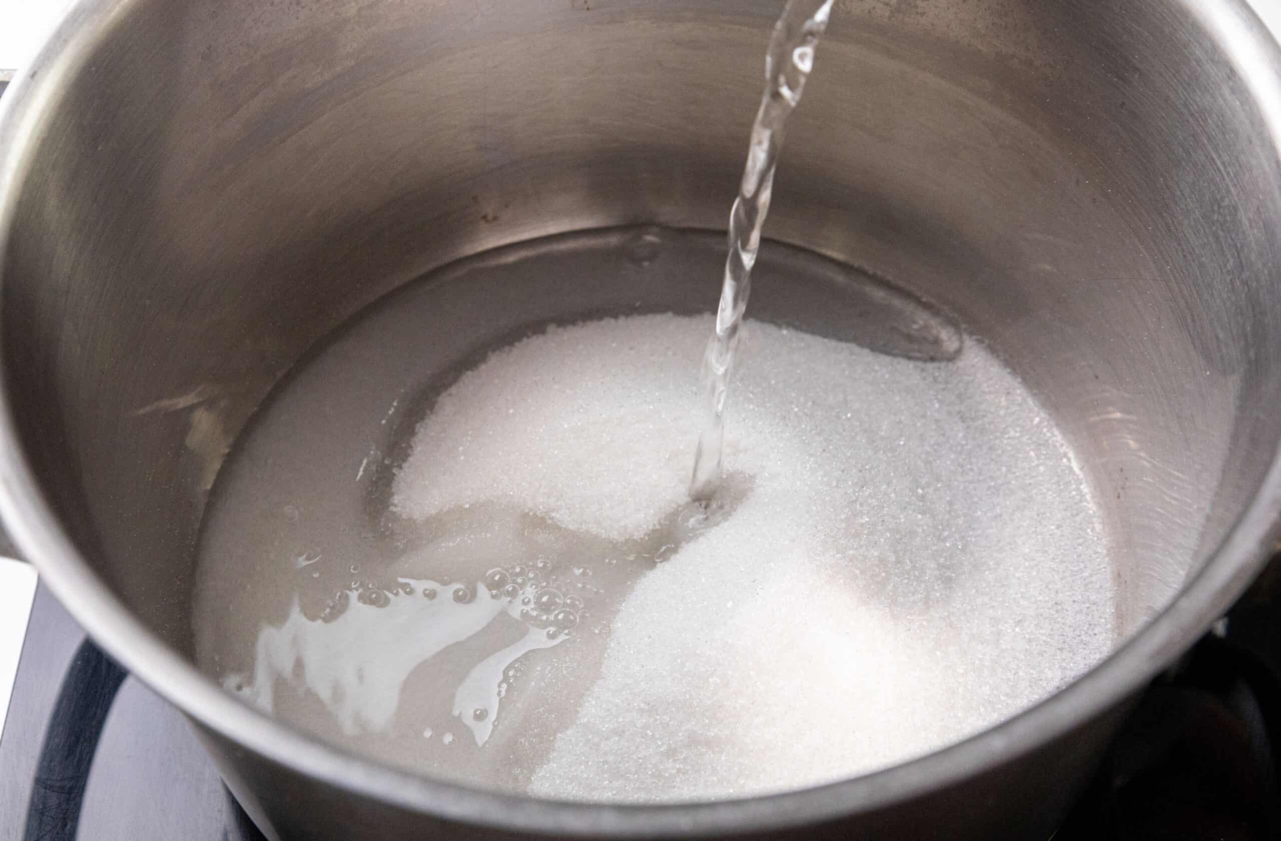Water being poured onto a mound of white sugar in a stainless steel pot.