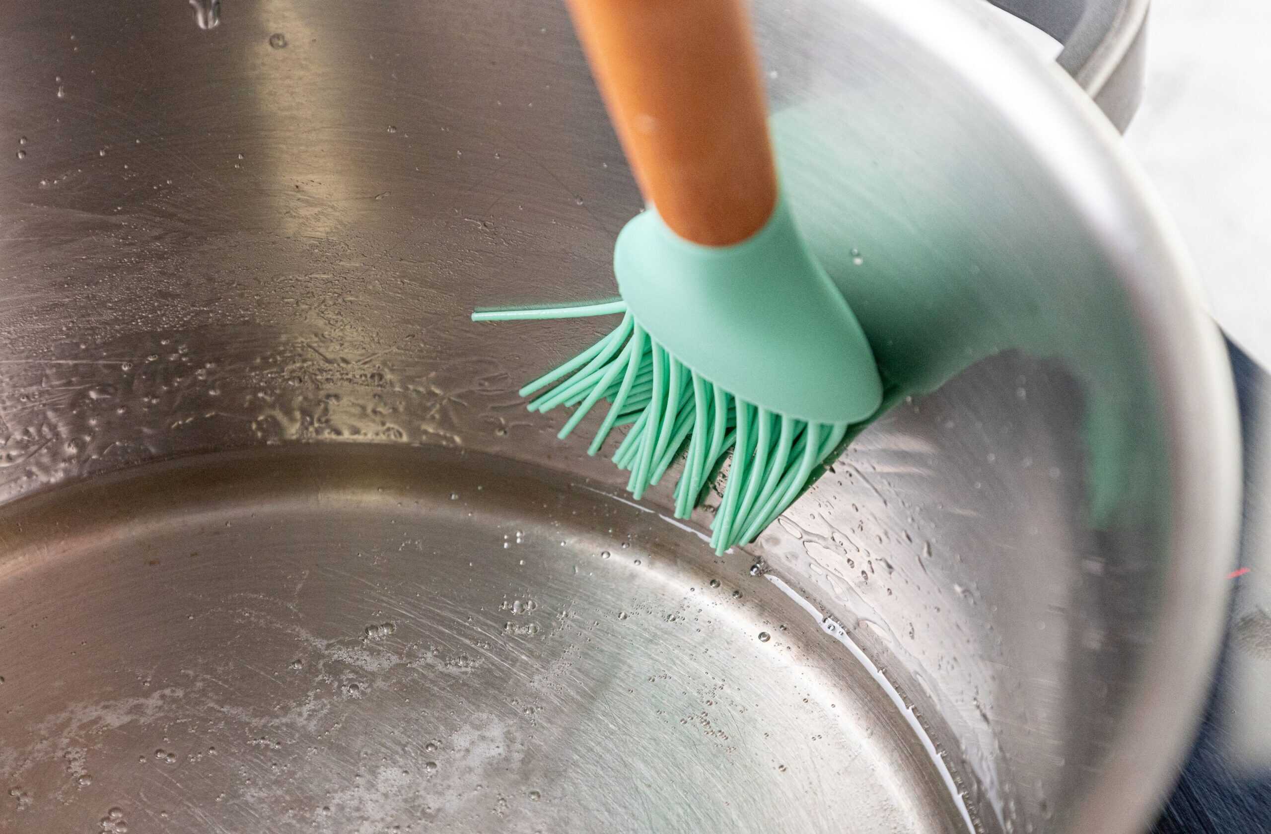 A green silicone brush with a wooden handle scrubs the inside of a stainless steel pot.