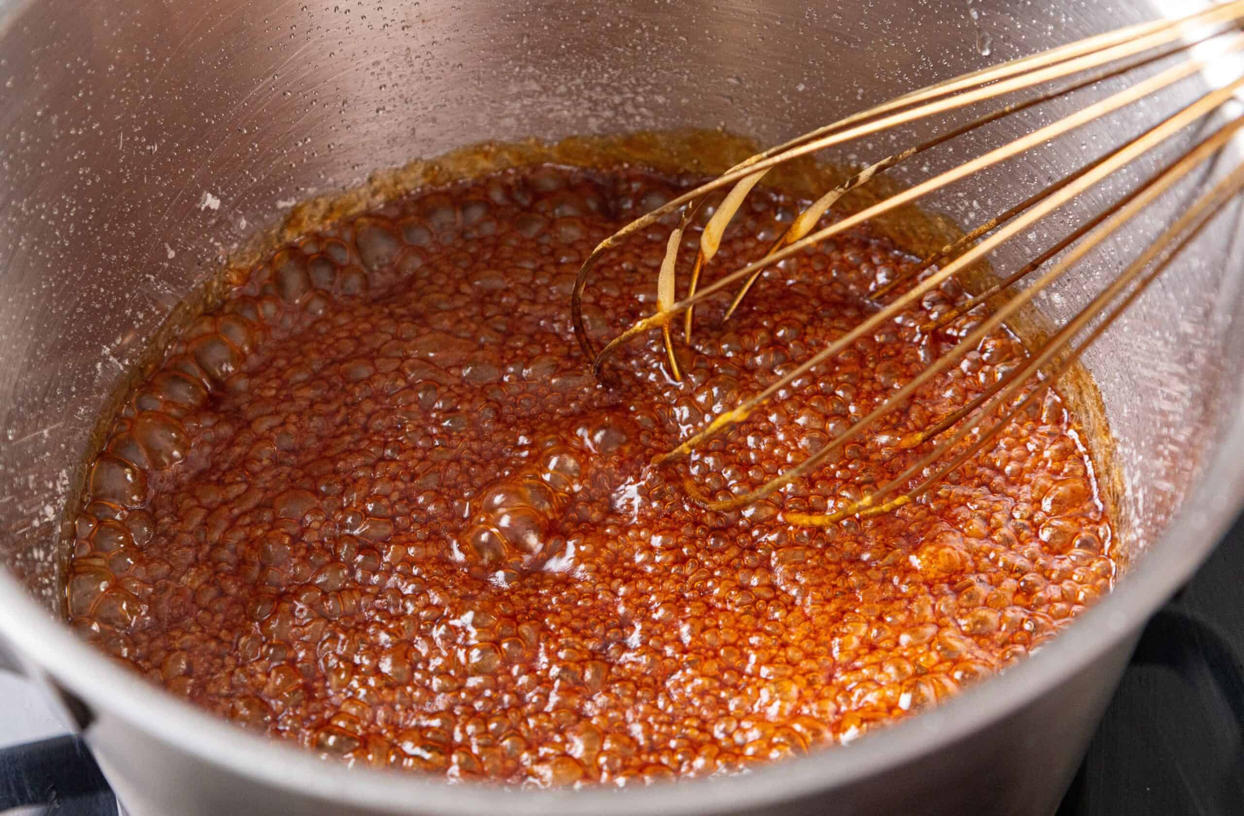 A metal whisk in bubbling caramel custard inside a stainless steel saucepan on a stovetop.