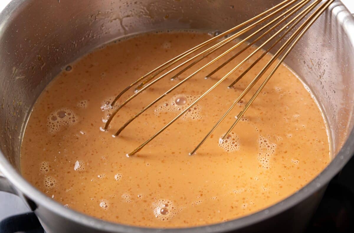 A metal whisk stirs light brown liquid in a stainless steel pot on a stovetop. The mixture bubbles gently.