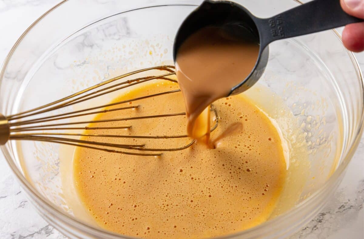 A measuring cup pours brown liquid into a glass bowl with yellow mixture, beside a metal whisk on a white countertop.