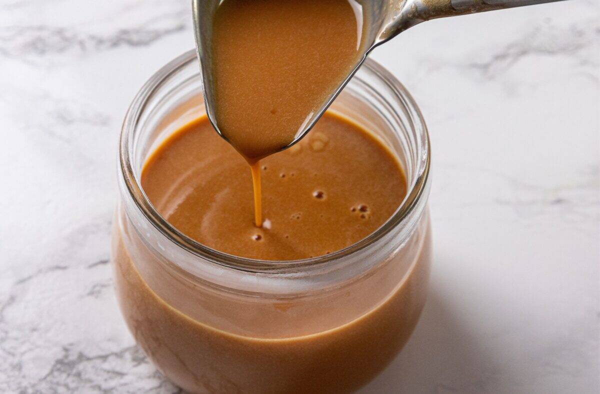 A spoon pouring caramel custard sauce into a glass jar already containing the same sauce, on a white marble surface.