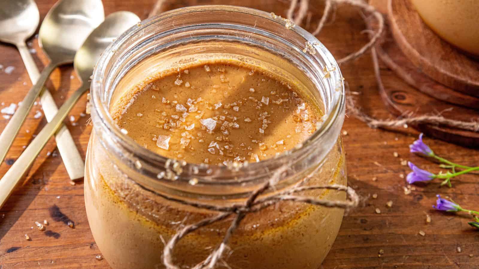 A glass jar of caramel custard topped with sea salt on a wooden table next to spoons and small purple flowers.
