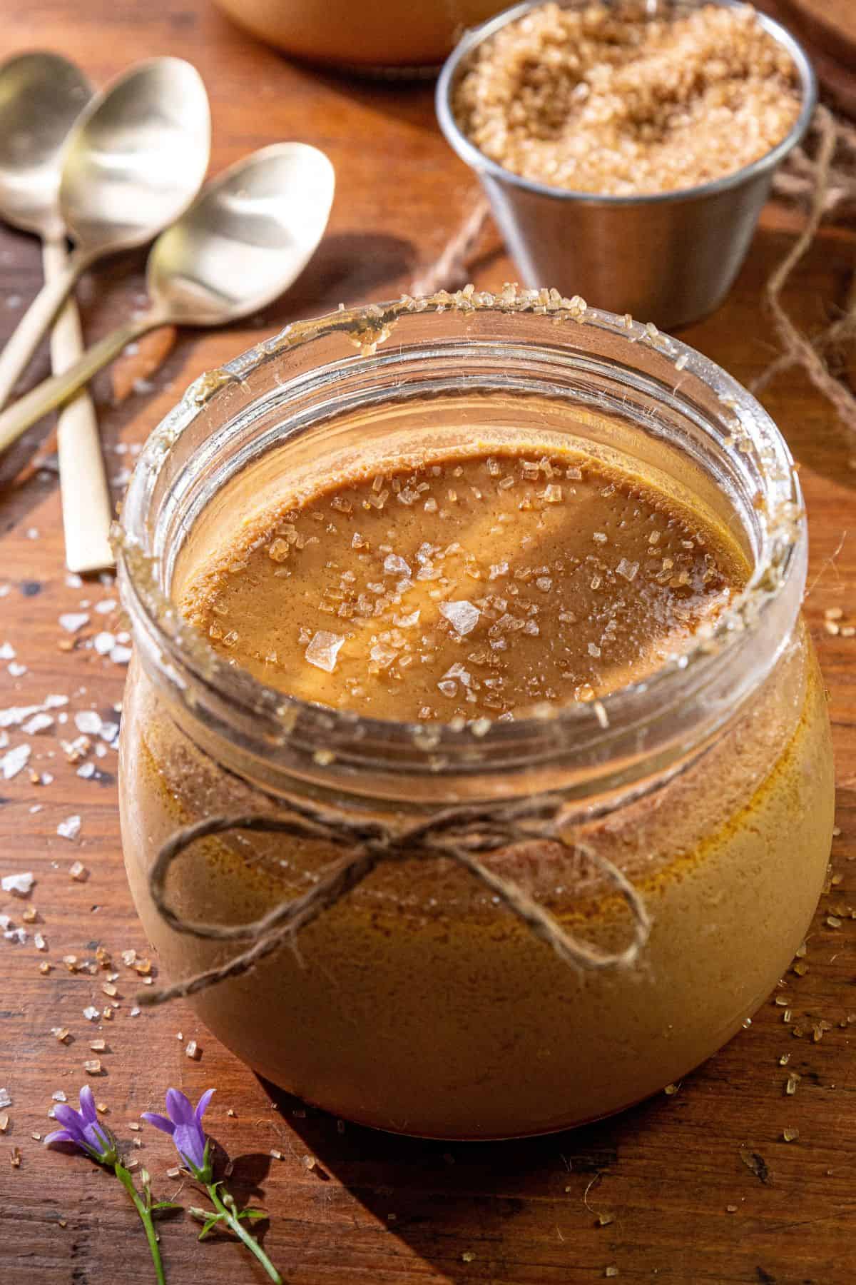 A glass jar of Salted Burnt Caramel Custard with coarse sea salt on top, three spoons, and a bowl of brown sugar on a wooden table.