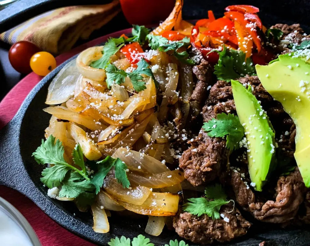 A close-up of a plate filled with sautéed onions, sliced colorful bell peppers, and seasoned beef, garnished with fresh avocado slices and cilantro. Shredded cheese is sprinkled on top. The vibrant dish is set against a dark background with hints of red and yellow accents.