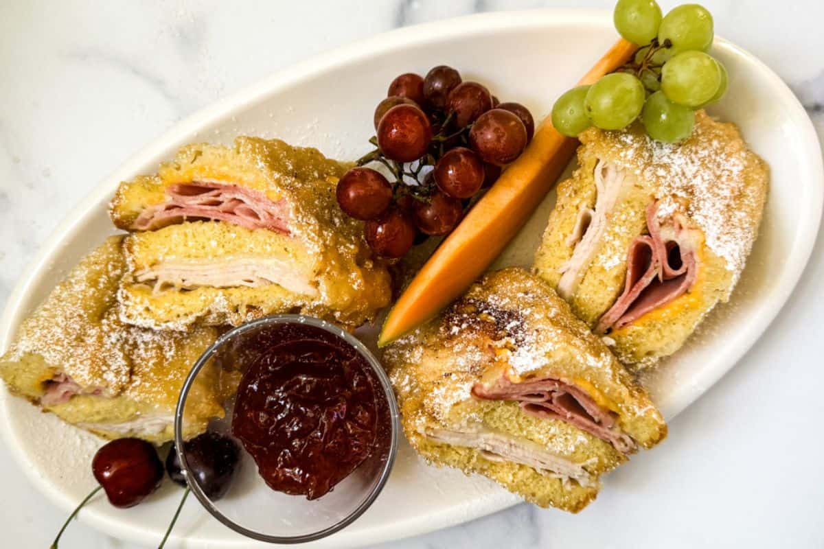 A plate of sandwiches dusted with powdered sugar, accompanied by grapes and a carrot stick, served with a side of jam.