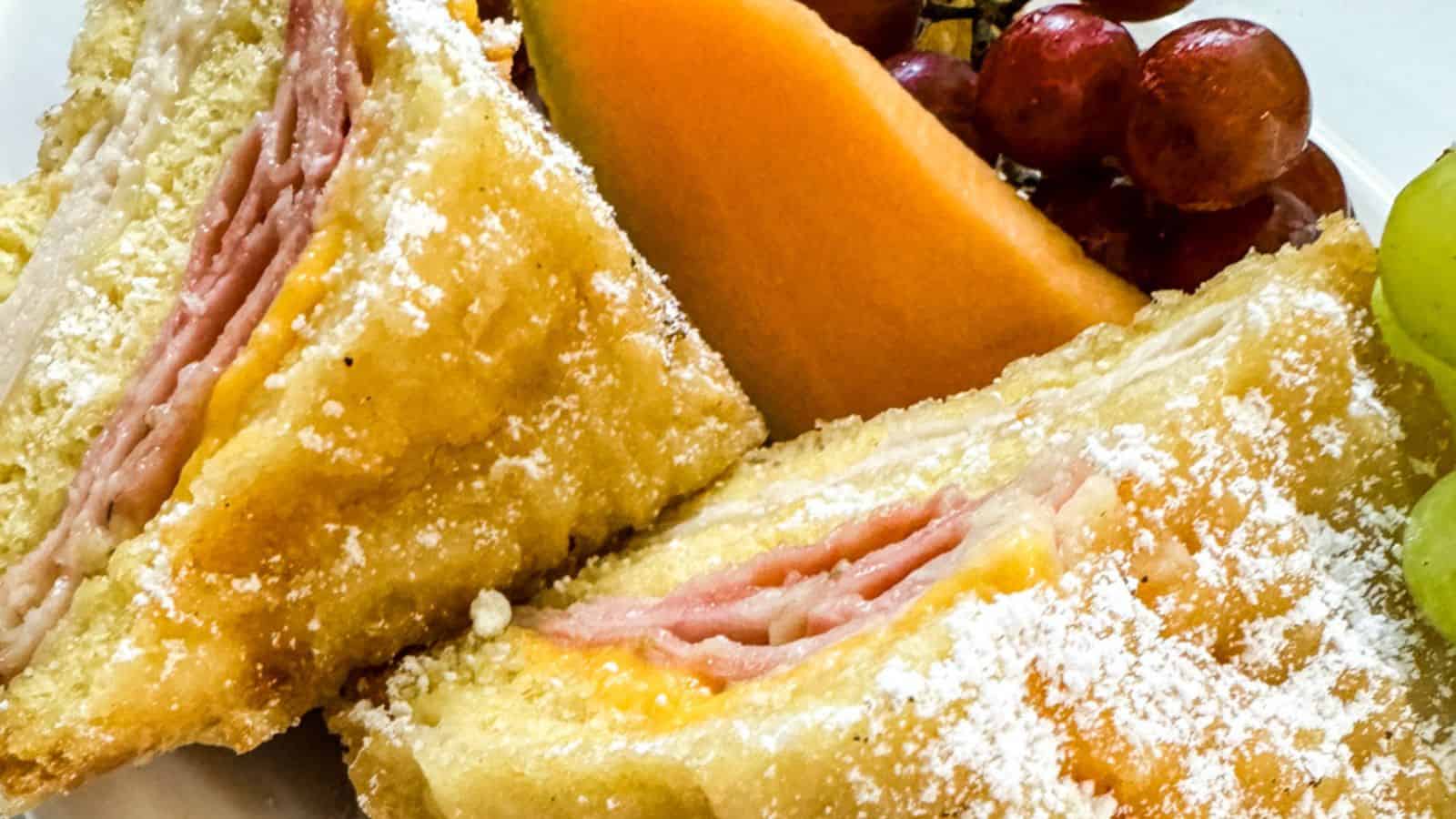 Two halves of a sandwich dusted with powdered sugar, served with cantaloupe slices and grapes.