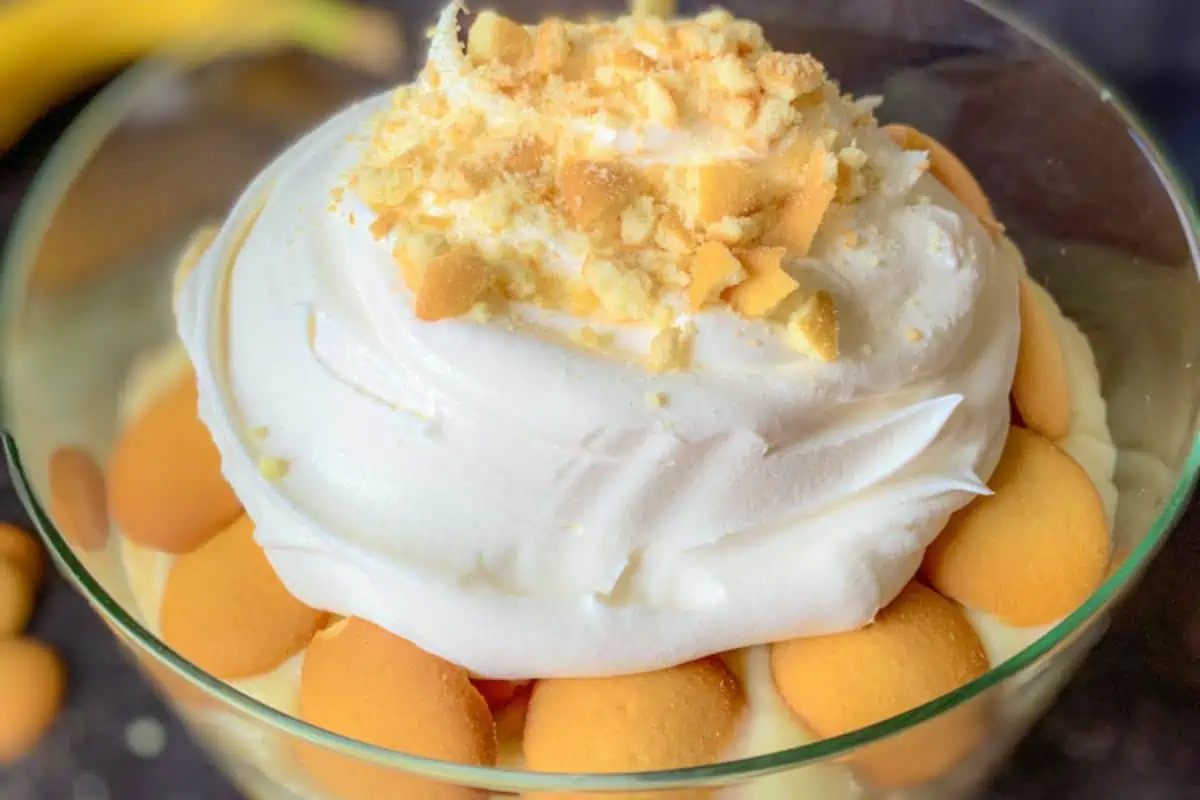 A close-up of a banana pudding trifle in a glass bowl. It has layers of creamy pudding, vanilla wafers, and is topped with a generous heap of whipped cream and crushed cookies. The dessert looks fluffy and indulgent with visible banana slices peeking through the layers.