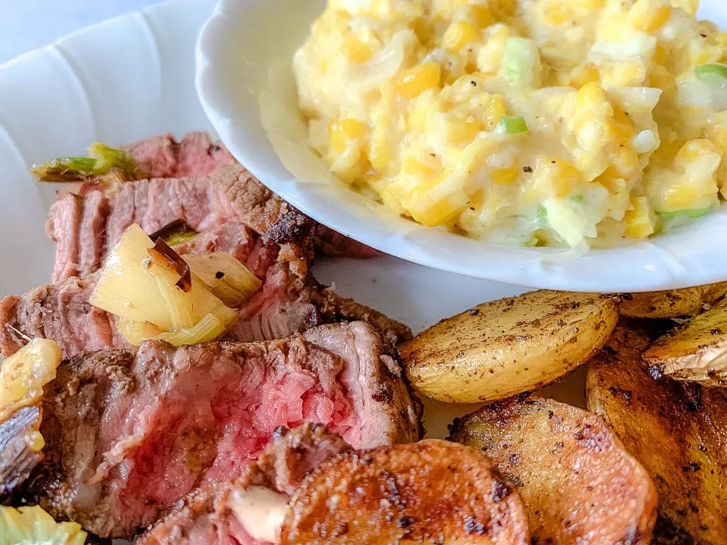 A plate featuring slices of medium-rare steak topped with grilled onions on the left, a bowl of creamy corn pudding on the top right, and golden-brown roasted potato slices on the bottom right. The food is arranged on a white plate.